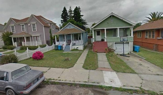A Google Street View screenshot showing a row of modest suburban houses with a small animal (possibly a cat) on the lawn of a gray house, with a silver car parked on the street.