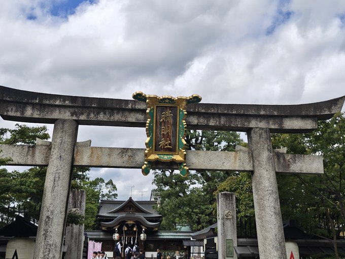 A traditional Japanese stone torii gate stands at the entrance to a Shinto shrine, featuring a central plaque with Japanese characters, with a cloudy sky and lush green trees in the background.