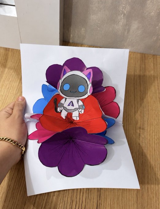 A hand holding a pop-up greeting card with a cute astronaut character surrounded by layered red, blue, and purple flower designs.