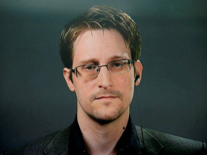The image features a man wearing glasses and a suit jacket with earphones plugged into his ears, giving him an intense look as he stares at the camera. The caption reads "EDWARD SNOWDEN SAYS, GOLD IS JUST BITCOIN THAT CAN'T BE SENT OVER THE INTERNET." This statement is likely related to the man's recent statement about Bitcoin and its limitations in terms of sending information over the internet.