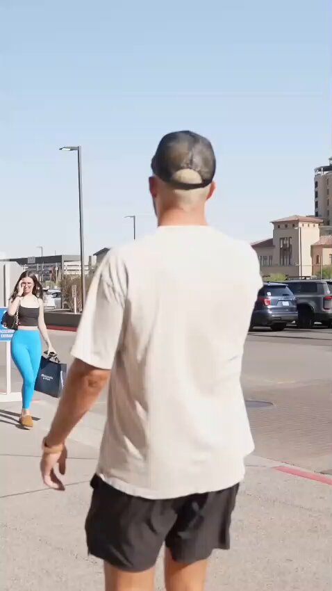 The image shows a man walking down a street while talking on his phone, wearing shorts and a hat. A woman is standing nearby with her handbag. The scene suggests that they are both engaged in separate activities or conversations.