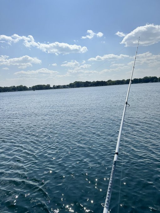 A view from a boat on a calm lake with a fishing rod extending into the foreground, a tree-lined shore in the distance, and a partly cloudy blue sky overhead.