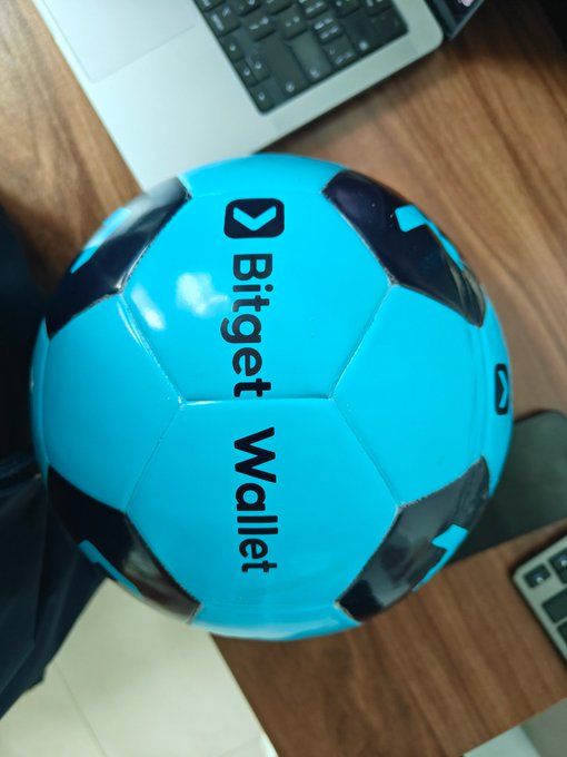 A small blue and black soccer ball branded with 'Bitget Wallet' sits on a wooden desk next to a laptop keyboard.