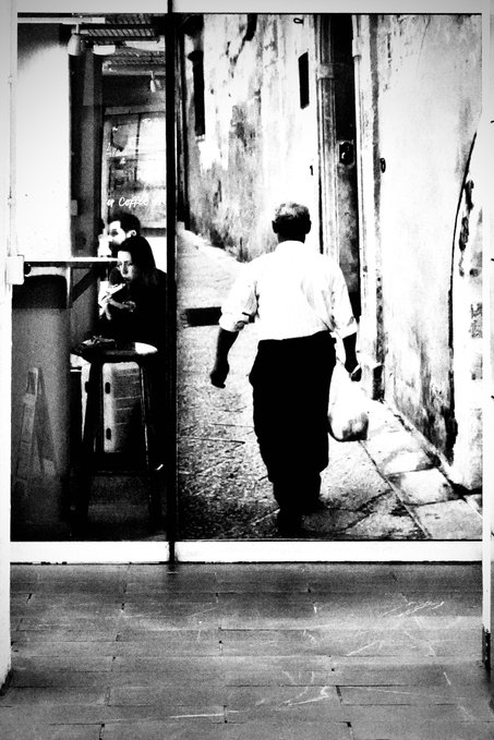 A black-and-white photograph of a large mural or photograph showing a man walking away down a narrow alley carrying a bag, with two people seated at a café on the left.