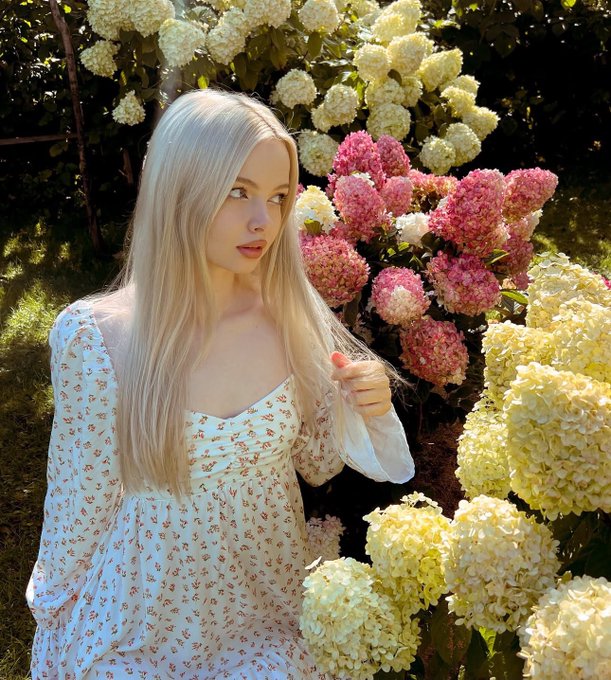 A blonde woman in a white floral dress stands next to blooming pink and white hydrangea bushes in a garden setting.