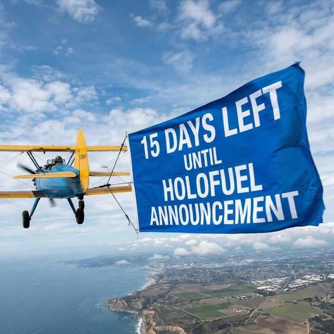 An aerial photograph of a yellow and blue biplane towing a banner reading '15 DAYS LEFT UNTIL HOLOFUEL ANNOUNCEMENT' over a coastal landscape.