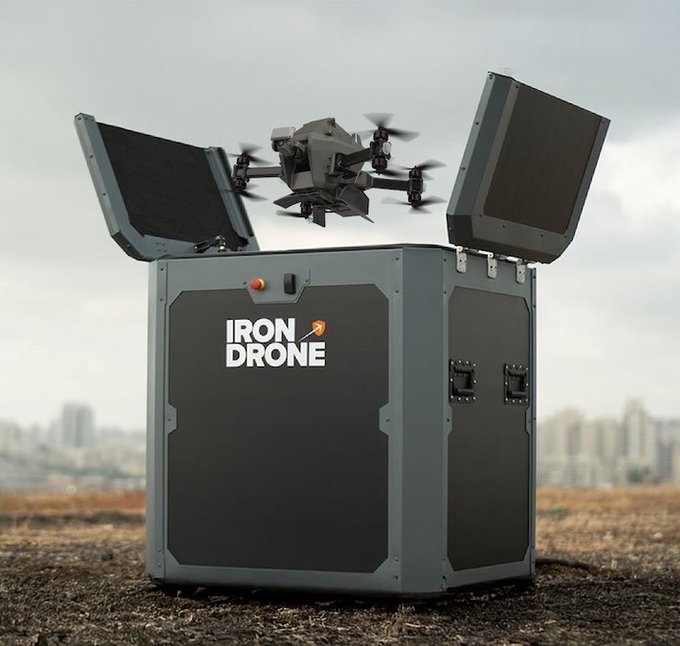 Render of a rugged gray case labeled 'IRON DRONE' with its lid open, revealing a quadcopter drone on top, set against a city skyline background.