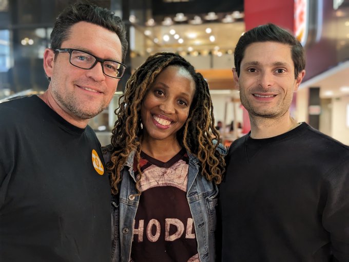 A selfie of three people smiling at the camera in what appears to be a bookstore; the middle woman has long dreadlocks and wears a denim jacket over a graphic tee, flanked by two men in dark shirts, one wearing glasses and a yellow button.