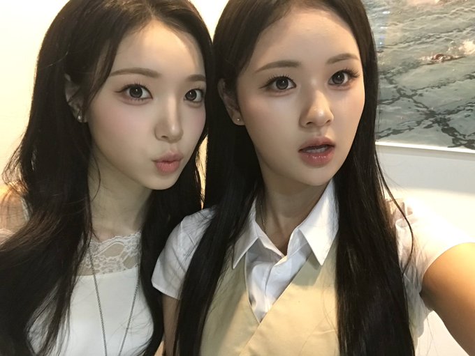 A close-up selfie of two young East Asian women, one making a kissing face and the other posing with slightly parted lips, both wearing white tops in what appears to be an indoor setting.