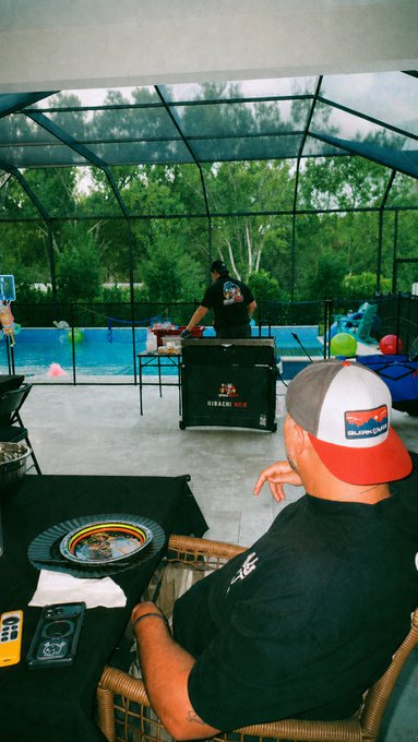 A man wearing a backward red, white, and grey cap sits under a screened patio enclosure, looking toward a cooking area with a Blackstone grill set and a pool visible in the background.