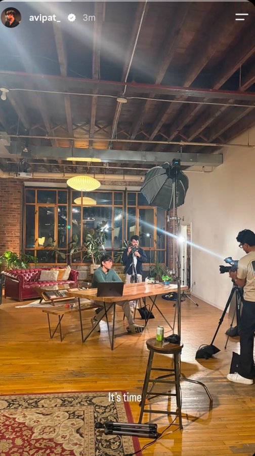 An Instagram story screenshot from 'avipat_' posted 3m ago shows a behind-the-scenes view of a video or photo shoot in a loft space with exposed wood beams, featuring a man at a wooden table and a camera operator with professional lighting equipment; the caption 'It's time' appears at the bottom.