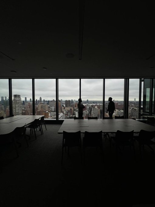A dark, silhouetted view of a modern high-rise conference room with two people standing by floor-to-ceiling windows, looking out at a cloudy city skyline.