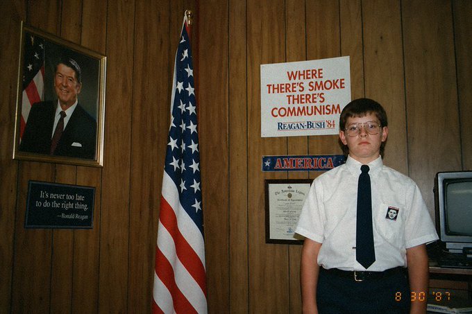 A young boy in a white shirt and black tie stands beside an American flag, with a portrait of Ronald Reagan and a "Reagan-Bush '84" sign reading "WHERE THERE'S SMOKE THERE'S COMMUNISM" displayed on the wood-paneled wall behind him. The dated photo (8/30/87) shows a pro-Reagan political display, humorously juxtaposed with the post's excitement about a speculative investment that will "sell out within a minute and 10x" — both capturing that timeless American optimism of betting big on something you believe in.