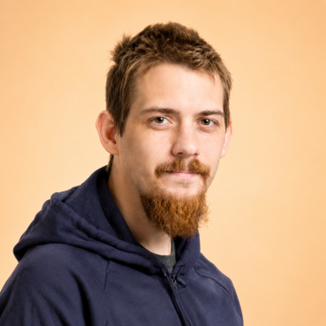 A portrait of a young man with reddish-brown hair and a goatee, wearing a navy blue hoodie against a tan background, representing a Bitcoin Core developer. The image accompanies news about Luke Dashjr's stance on Bitcoin's proof-of-work and the ongoing debates surrounding BIP 110.