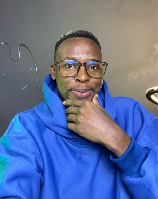 A close-up selfie of a man wearing glasses and a blue hoodie, posing thoughtfully with his hand on his chin against a dark gray wall.