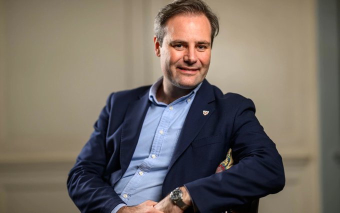 The image shows a man in a navy blazer and light blue shirt smiling at the camera, which appears to be a professional portrait of crypto billionaire Ben Delo, the donor featured in the post about his £36m donation to Reform UK.