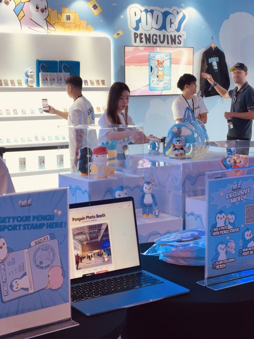 A Pudgy Penguins merchandise booth at what appears to be a convention, featuring staff in white shirts, a photo booth laptop, exclusive merch displays, and signage reading 'PUDGY PENGUINS'.