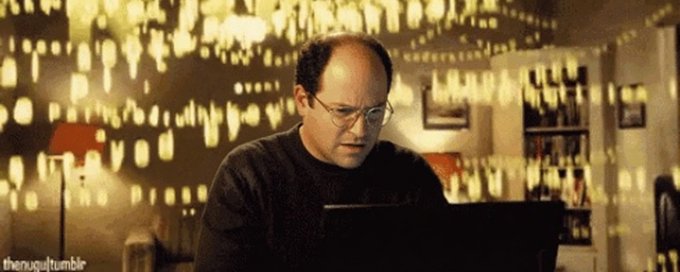 A man with glasses, resembling George Costanza from Seinfeld, stares intensely at a laptop screen in a warmly lit room with bookshelves.
