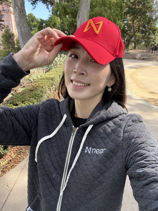 A woman taking a selfie wearing a red "N" cap and a dark gray "Mnear" zip-up hoodie in a park setting.