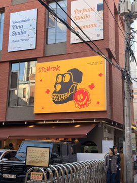 A street view of a building featuring a large orange billboard with a black skull graphic and red 'STORM' text, alongside signs for 'Miyawaki Ding Studio' and 'Perfume Studio' above.