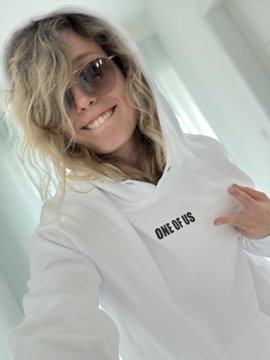 A smiling blonde woman wearing sunglasses and a white hoodie that reads "ONE OF US" takes a selfie, aligning with the caption's message about believing in magic and tagging @CryptoWizardd and @NonPlayableCoin, likely promoting community membership in a crypto project.