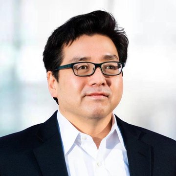 A professional headshot of an Asian man with dark hair, wearing black-rimmed glasses and a dark suit jacket over a white shirt.