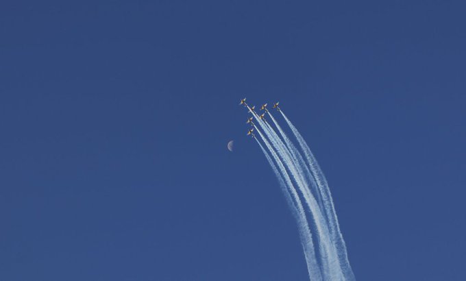A formation of jets flies in a V-shape across a deep blue sky, leaving long white contrails behind them with the moon visible in the background.
