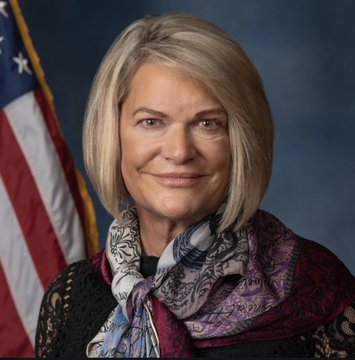 The image features a woman with blonde hair wearing a black dress and a colorful scarf around her neck. She has a smile on her face as she poses for the picture. The background of the photo includes an American flag, which adds a patriotic touch to the scene. The caption "Senator Cynthia Lummis, today: ?" suggests that this image might be related to a post about Bitcoin or cryptocurrency.