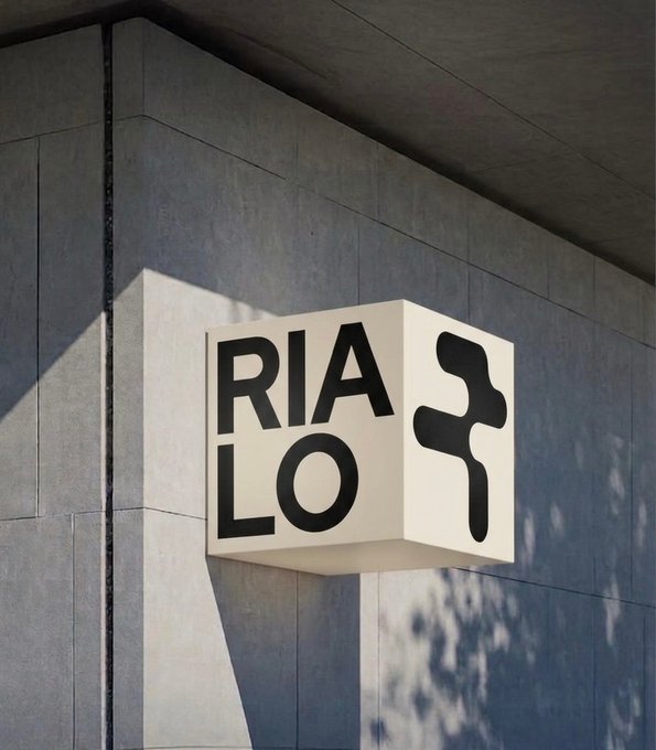 A white cube-shaped sign mounted on a grey tiled wall displays the word "RIALO" in bold black letters on one face and a stylized black "R" logo on another.