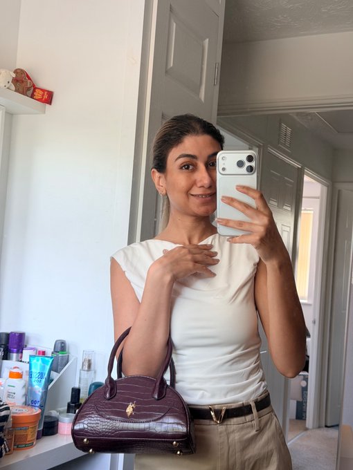 A woman takes a mirror selfie in what appears to be a bathroom, wearing a white sleeveless top and holding a burgundy crocodile-textured handbag with a gold logo.