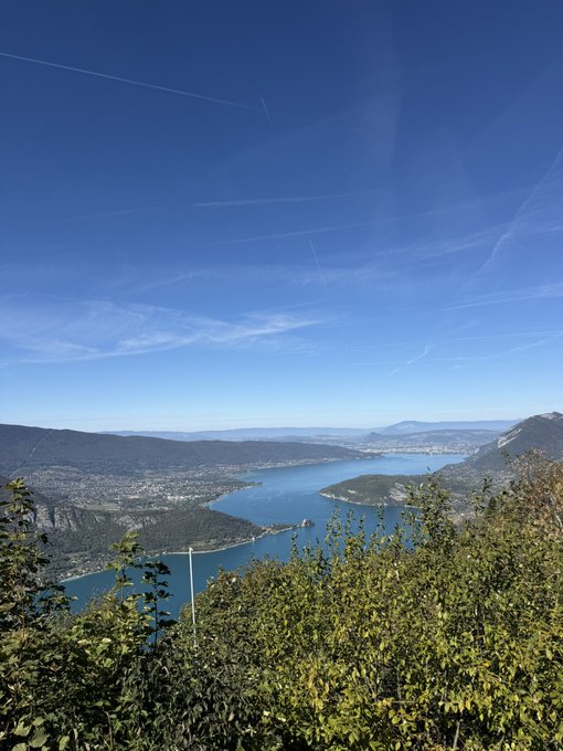 A scenic landscape view of a blue lake surrounded by mountains and forest, taken from an elevated vantage point under a clear sky with visible contrails.
