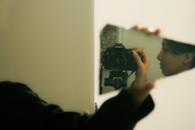 A person taking a mirror selfie with a Canon DSLR camera, their face partially visible in the reflection within a fragmented or torn frame effect.