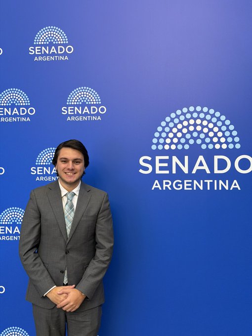 A man in a gray suit and striped tie smiles while standing in front of a blue backdrop with the logo 'SENADO ARGENTINA' repeated.