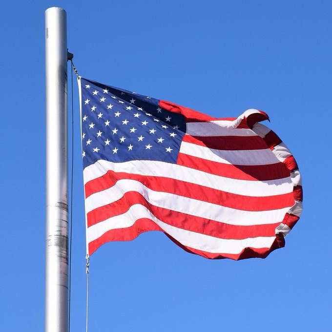 An American flag waves against a clear blue sky, symbolizing national unity as 39 U.S. state banking associations come together under the "BankChain Alliance" to launch a nationwide blockchain network for regulated banks. The flag's patriotic imagery aligns with the post's message of unified, country-wide collaboration in blockchain innovation.