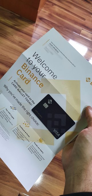 A hand holding a 'Welcome to your Binance Card' promotional flyer featuring a black Visa debit card and listing benefits like Paying Coins, Earning & Saving, and Travel VIP Card over a wooden floor background.