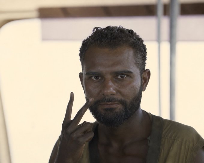A close-up portrait of a bearded man with short curly hair making a gesture pointing to his eye, wearing a tan shirt and standing under what appears to be a canopy.