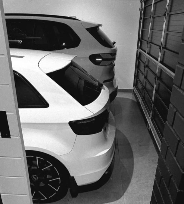 A black and white photo of two cars parked side by side in a narrow garage next to a brick wall, with the rear of a white hatchback prominent in the foreground.