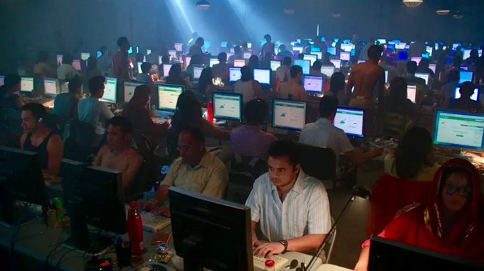 A crowded LAN party or gaming event with dozens of people seated at rows of computers, illuminated by the glow of monitors in a dimly lit hall.