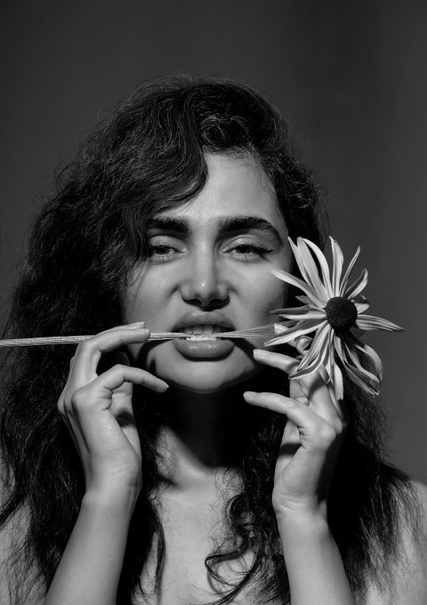 A black-and-white portrait of a woman with long dark hair holding a daisy horizontally in front of her lips with both hands.