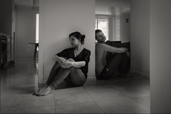A black and white photograph showing a young woman and man sitting on the floor, leaning against opposite sides of a hallway partition, looking in different directions.