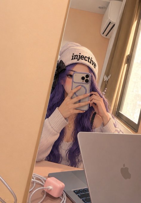 A mirror selfie of a person with long purple hair wearing a white beanie with the word 'injective' printed on it, holding an iPhone, with a MacBook and a pink charger visible on the desk.