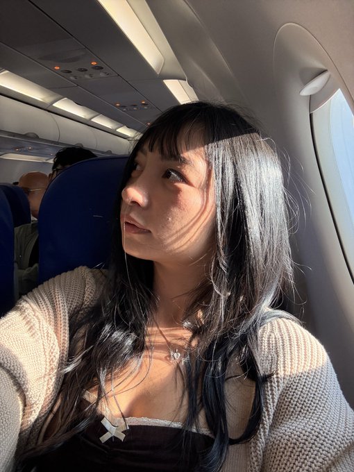 Selfie of a young woman with long dark hair sitting in an airplane window seat, wearing a cream-colored cardigan over a dark top with a butterfly necklace.