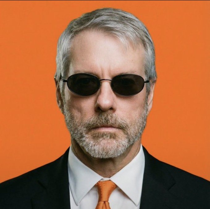 A middle-aged man with gray hair and a salt-and-pepper beard wears dark oval sunglasses, a black suit, white dress shirt, and an orange tie, posing against a vibrant orange background that matches the bullish excitement of the post announcing $MSTR's return to the top 500 companies of the world, with the upward rocketing emojis mirroring his confident, forward-facing stance symbolizing the company's triumphant climb back into elite global rankings.