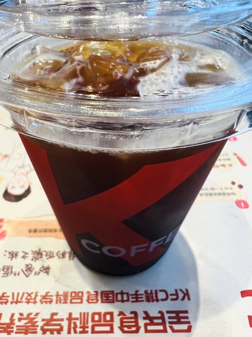 A close-up shows a clear plastic cup filled with iced coffee, bearing a large 'K' logo and the word 'COFFEE', sitting on a tray with Japanese text.