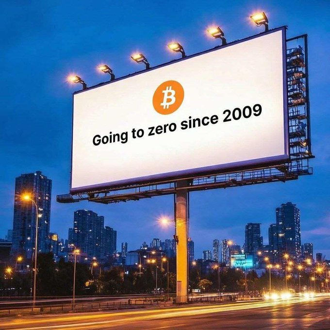 A large billboard at dusk displaying the Bitcoin logo with the text 'Going to zero since 2009' set against a city skyline.