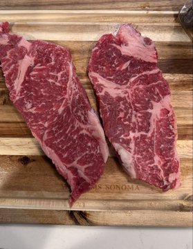 Two raw cuts of marbled beef, likely New York strip or similar steaks, rest on a wooden cutting board.