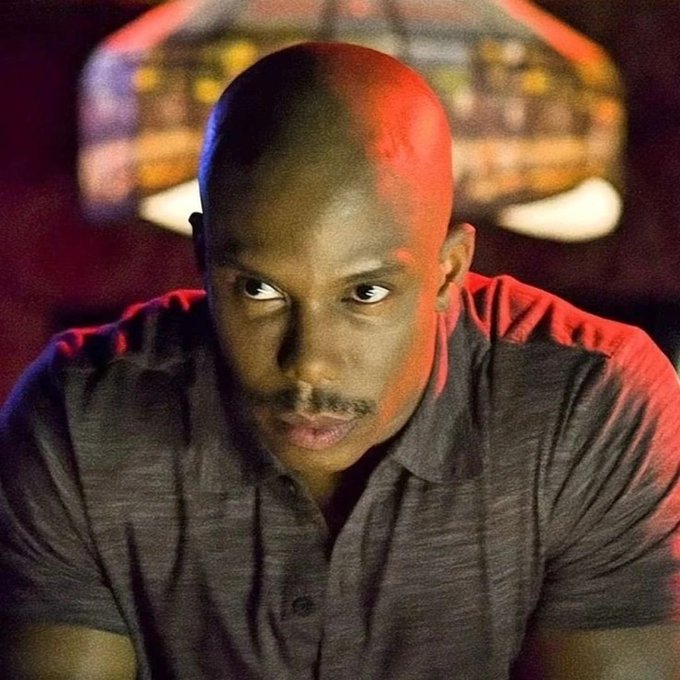 A dramatic portrait of a bald man with a mustache wearing a dark polo shirt, illuminated by red and blue lighting with a blurred arcade-style display in the background.