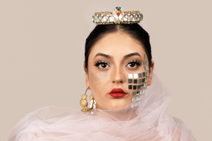 A close-up portrait of a woman wearing a jeweled crown and red lipstick, with silver mosaic tiles covering one side of her face and gold circular makeup on the other cheek, against a beige background.