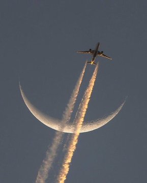 An airplane flies away from the viewer, leaving twin contrails, positioned above a thin crescent moon in a deep blue sky.