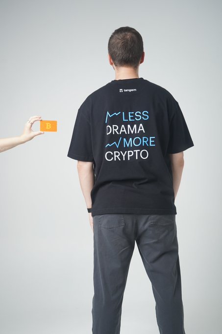 A man seen from behind wearing a black Tangem t-shirt with the slogan 'LESS DRAMA MORE CRYPTO', while a hand offers him a Bitcoin coin.